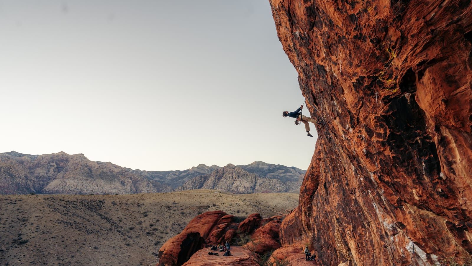 A climber suspended on a red sandstone wall above desert mountains.